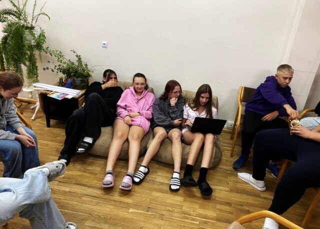 Młodzież siedząca wewnątrz pomieszczenia. Jedna osoba trzyma na kolanach laptop.