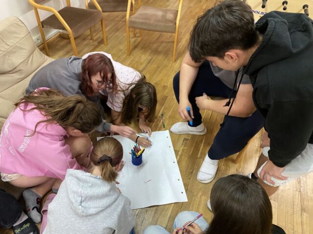 Grupa młodzieży pracuje w skupieniu nad rysunkiem rozłożonym na podłodze.