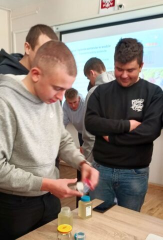 Uczniowie w sali wykłądowej, przy stolikach, prowadzą badania próbek wody. W tle ekran z wyświetlaną prezentacją.