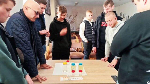 Grupa uczniów wraz z opiekunem. Stoją przy stoliku na którym są próbki wody oraz tablice do odczytów.
