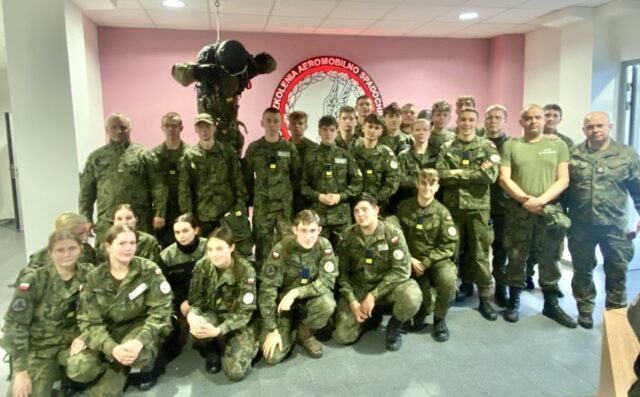 Grupa kadetów klasy OPW wraz z opiekunami w umundurowaniu wewnątrz budynku. W tle logo Ośrodka Szkolenia Aeromobilno Spadochronowego oraz manekin żołnierza.