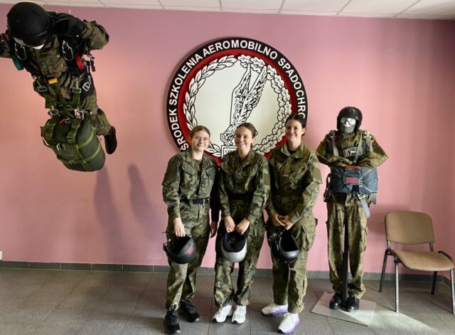 3 kadetki klasy OPW wewnątrz pomieszczenia. W tle logo Ośrodka Szkolenia Aeromobilno Spadochronowego oraz dwa manekiny żołnierzy.