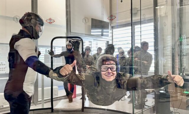 Kadet klasy OPW wraz z instruktorem w tunelu aerodynamicznym. Na zewnątrz tunelu inni uczestnicy szkolenia.