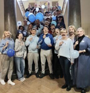 Grupa  młodzieży wraz z opiekunami na schodach w budynku szkolnym. Większość osób nosi ubrania w kolorze niebieskim oraz przykłada do ust wąsy, pokazując w ten sposób wsparcie akcji Movember. Widoczne akże balony w kolorze niebieskim. Osoby na zdjęciu 