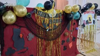andrzejki_internat4 Dekoracja andrzejkowa. Na czerwonym tle napis w kolorach czarnym i złotym Andrzejki 2025, kolorowe balony i symbole zabawy andrzejkowej.