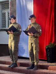 apel_odzyskanie_niepodleglosci01 Dwóch uczniów w umundurowaniu i nakryciach głowy na schodach przed wejściem do budynku szkoły. Jeden z nich trzyma w ręku mikrofon. Patrzą na trzymane w rękach teczki. W tle rozwieszona flaga Polski.