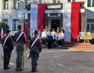 apel_odzyskanie_niepodleglosci13 Grupa młodzieży w strojach galowych na schodach przed wejściem do budynku szkoły. W tle rozwieszone flagi Polski. Na pierwszym planie poczet sztandarowy.