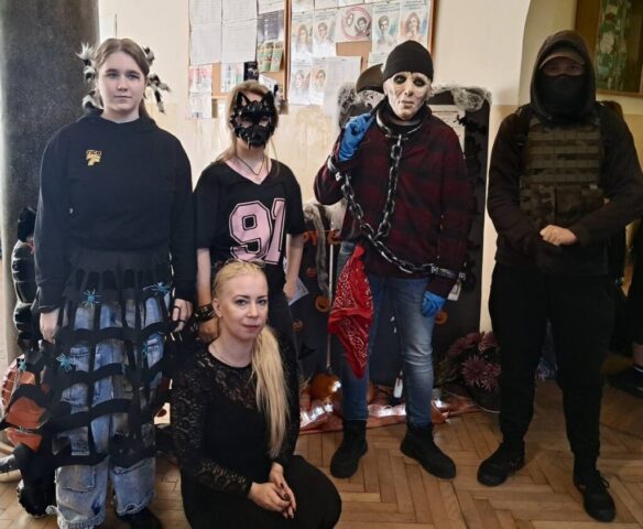 4 osoby w przebraniu i 1 kobieta w czarnym stronu wewnątrz, prawdopodobnie korytarz szkolny. W tle kolumna, gazetka ścienna oraz fragment dekoracji na Halloween.