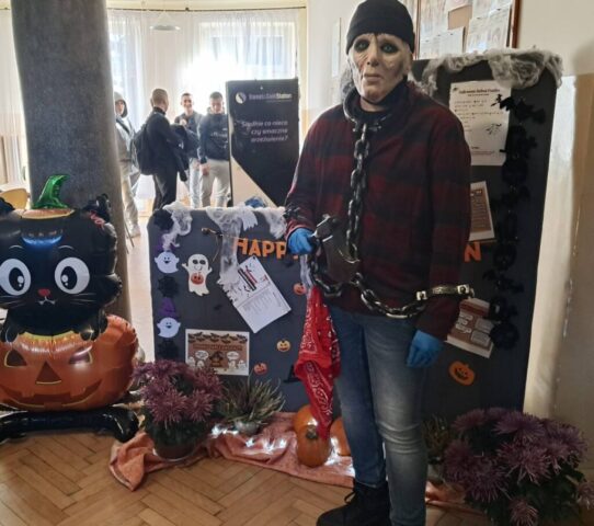 Osoba w przebraniu pozuje do zdjęcia na tle gazetki tematycznej z okazji Halloween. Udekorowany korytarz szkolny. W tle grupka uczniów.
