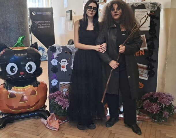 Dwie uczennice w przebraniu z okazji Halloween. Stoją na tle dekoracji. Po lewej stronie balony w kształcie dyni oraz czarnego kota.