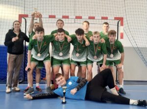 Grupa młodzieży w strojach sportowych wraz z opiekunem. Stoją na tle bramki w hali i prezentują medale, które mają na szyjach. Jedna osoba trzyma w rękach puchar. Jedna osoba leży na podłodze, przed nią jest kolejny puchar. 