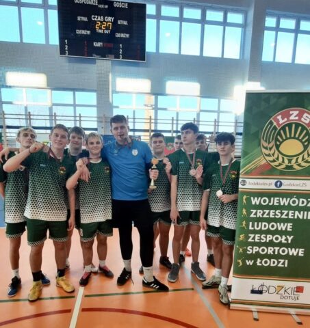 Drużyna sportowa prezentująca zdobyte medale i puchar. Po prawej stronie banner reklamowy LZS oraz w tle zegar sportowy oraz tablica wyników zawieszone nad drabinkami.