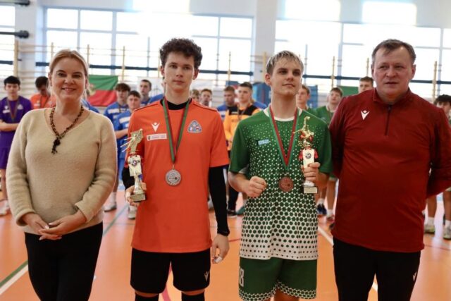 Zawodnicy z medalami na szyjach i pucharami w dłonie oraz dwie osoby dorosłe. Stoją w hali sportowej. W tle inne drużyny sportowe.