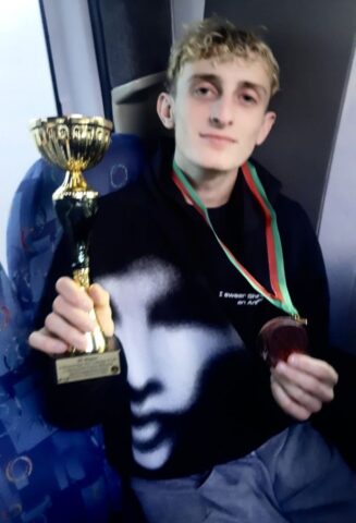 Młody mężczyzna siedzi i prezentuje medal oraz puchar.
