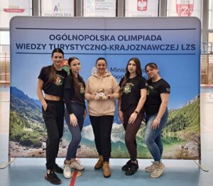4 uczennice w czarnych koszulkach z wizerunkiem misia i napisem Zakopane 2025 wraz z opiekunem stoją na tle banneru reklamującego Ogólnopolską Olimpiadę Wiedzy Turystyczno - Krajoznawczej LZS. Osoba stojąca w środku trzyma w ręku maskotkę. 