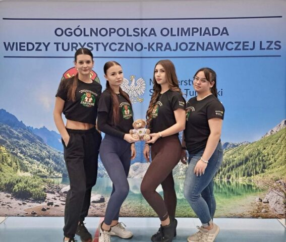 4 uczennice w czarnych koszulkach promocyjnych z wizerunkiem niedźwiedzia i napisem Zakopane 2025 stoją na tle banneru reklamowego. W ręku trzymają maskotkę Olimpiady.