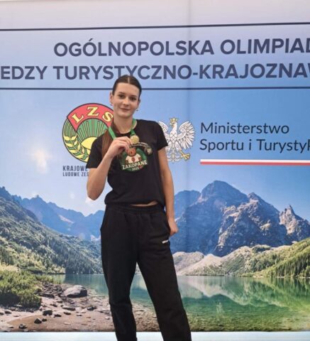 Młoda kobieta w czarnej koszulce z wizerunkiem niedźwiedzia i napisem Zakopane 2025, mająca na szyi medal na zielonej szarfie, stoi na tle banneru reklamującego wydarzenie - Ogólnopolska Olimpiada Wiedzy Turystyczno- Krajoznawczej. Na banerze widok Gór i jeziora, logo LZS, Orzeł w koronie oraz napis Ministerstwo Sportu i Turystyki.