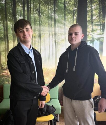 Dwóch młodych mężczyzn stoi w pomieszczeniu na tle fototapety przedstawiającej las w słoneczny dzień. Mężczyźni ściskają sobie dłonie w geście gratulacji.