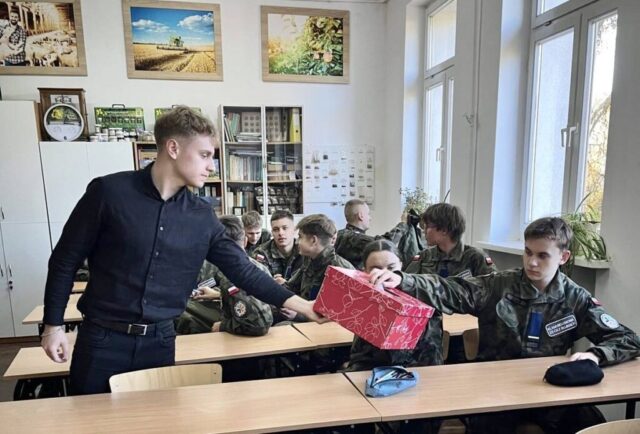 Grupa uczniów w umundurowaniu klasy wojskowej w sali lekcyjnej. Siedzą wławkach. Na pierwszym tle młody mężczyzna ubrany na czarno trzyma w ręku pudełko, do którego jeden z uczniów wrzuca karteczkę.