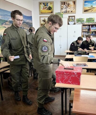 Uczniowie w klasie lekcyjnej. Na pierwszym planie ławka, na której stoi pudełko. Po lewej stronie dwóch uczniów, jeden wrzuca karteczkę do pudełka, drugi czeka za nim na swoją kolej. W tle inni uczniowie siedzący w ławkach.