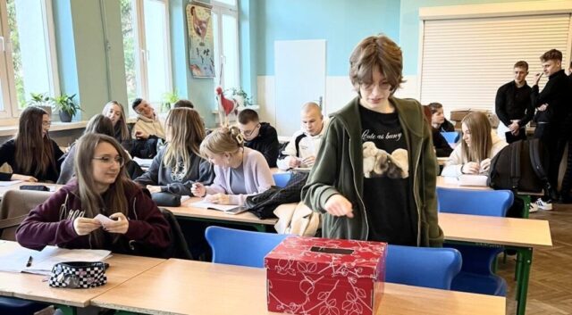 Uczniowie w klasie lekcyjnej. Na pierwszym planie ławka, na której stoi pudełko. Jeden z uczniów wrzuca karteczkę do pudełka, kolejna uczennica trzyma karteczkę w ręku i czeka na swoją kolej. W tle inni uczniowie siedzący w ławkach lub stojący w tyle klasy.