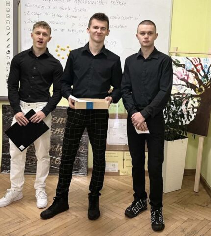 Trzech uczniów w sali lekcyjnej. Stoją przed tablicą, trzymają w ręku pudełko, teczkę i plik karteczek.