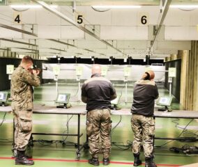 zawody_strzeleckie02 3 osoby w umundurowaniu wewnątrz strzelnicy. Stoją przy stanowiskach 4, 5 i 6 i oddają strzały.