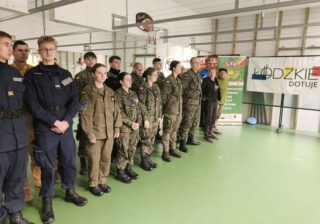 zawody_strzeleckie05 Grupa młodzieży w umundurowaniu stoi w dwuszeregu na sali gimnastycznej. W tle widoczne banery LZS i łódzkie dotuje.