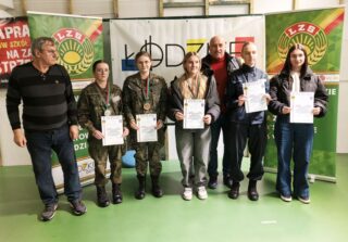 zawody_strzeleckie06 5 dziewcząt oraz 2 instruktorów wewnątrz sali. Dziewczęta trzymają w rękach dyplomy, na szyjach mają medale. W tle banery reklamowe organizatorów zawodów strzeleckich..