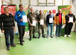 zawody_strzeleckie08 3 Chłopców oraz 4 instruktorów wewnątrz sali. Chłopcy trzymają w rękach dyplomy, na szyjach mają medale. W tle banery reklamowe organizatorów zawodów strzeleckich..