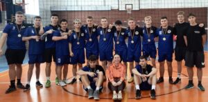 Grupa 15 młodych zawodników sportowych w strojach granatowych i czarnych wraz z instruktorem. Stoją i siedzą w hali sportowej. 