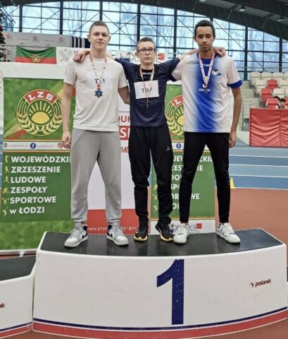 3 uczniów w strojach sportowych stoi na podium w dużej hali sportowej. Na szyjach mają medale. W tle trybuny oraz bannery reklamowe LZS.