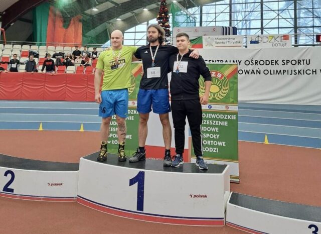 3 Uczniów w strojach sportowych stoi na podium w dużej hali sportowej. Na szyjach mają medale. W tle trybuny oraz kibice oraz bannery reklamowe LZS, COS, Ministerstwa Sportu i Turystyki oraz Łódzkie dotuje.