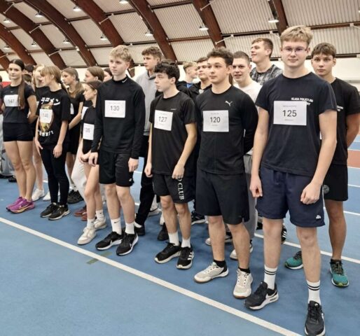 Grupa dziewcząt i chłopców w strojach sportowych z numerami startowymi w dużej hali sportowej.