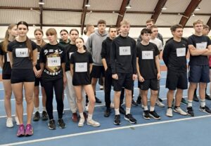 Grupa młodzieży w strojach sportowych z numerami startowymi w dużej hali sportowej. 