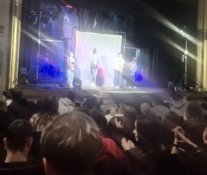 4 młodych ludzi na scenie. Na pierwszym planie publiczność zgromadzona na spektaklu teatralnym. 