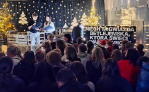 Sala gimnastyczna udekorowana na okoliczność Bożego Narodzenia. Dwóch uczniów, prowadzących przegląd pastorałek i zgromadzona publiczność.