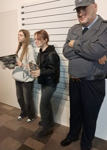 Dwie kobiety w muzeum z figurą woskową bohatera serialu Ojciec Mateusz. Sceneria z komisariatu policji.