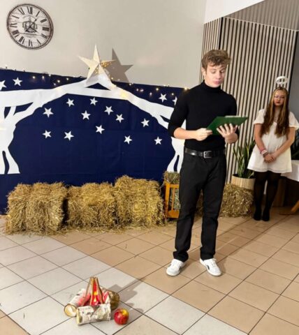 wigilia_internat02 Dwie młode osoby w sali przystrojonej na okoliczność Jasełek. Mężczyzna czyta coś, po prawej stronie kobieta w stroju Anioła.