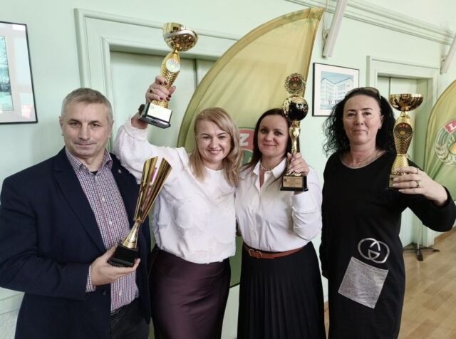 lzs_podsumowanie1 4 osoby (3 kobiety i mężczyzna wewnątrz pomieszczenia. Trzymają w rękach trofea sportowe. W tle bannery LZS.