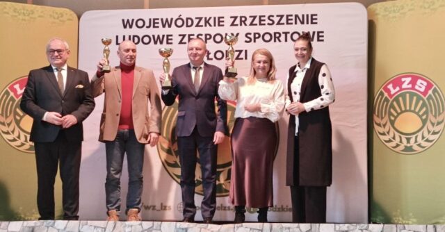 lzs_podsumowanie3 2 kobiety i 3 mężczyzn stoją na scenie. 3 osoby trzymają w ręku trofea sportowe. W tle baner reklamowy i logo LZS