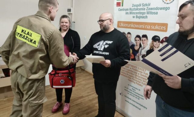 Uczeń klasy strażackiej w umundurowaniu odbiera z rąk organizatorów dyplom. W tle baner reklamowy ZS CKZ im. Wincentego Witosa w Bujnach.