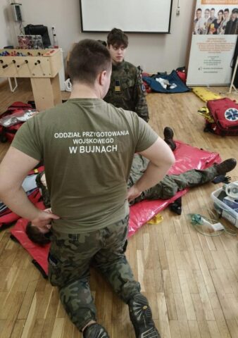 Uczestnicy kursu KPP podczas szkolenia z udzielania pierwszej pomocy. Widoczny sprzęt medyczny i plecak ratowniczy.