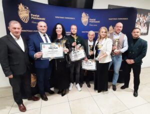 8 osób stojących na tle banerów reklamowych Starostwa Powiatu Piotrkowskiego. Część osób trzyma w rękach dyplomy i trofea sportowe. 