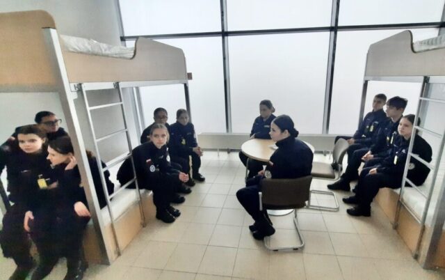 Uczniowie klasy mundurowej wewnątrz pomieszczenia. Widoczne dwa łózka piętrowe i stolik z krzesłami.