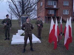 3 uczniów klasy opw w umundurowaniu stoi przy obelisku. Po prawej stronie stojak z flagami Polski.