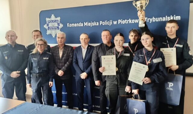 Zwycięska drużyna uczniów wraz z organizatorami. Pozują na tle baneru reklamowego Komendy Miejskiej Policji w Piotrkowie Trybunalskim. Uczniowie trzymają w ręku puchar i dyplomy, na szyjach mają medale.
