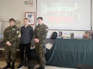 3 uczniów stoi w pomieszczeniu. W tle slajd promujący konkurs. widoczna też wystawa tematyczna. 