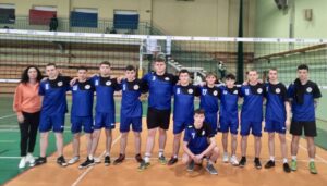 Drużyna 12 zawodników w granatowych strojach sportowych wraz z opiekunem w sali gimnastycznej na tle boiska do siatkówki. 