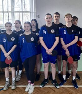 Grupa młodych zawodników w jednolitych strojach sportowych stoi w sali gimnastycznej, trzymając rakietki do tenisa stołowego.